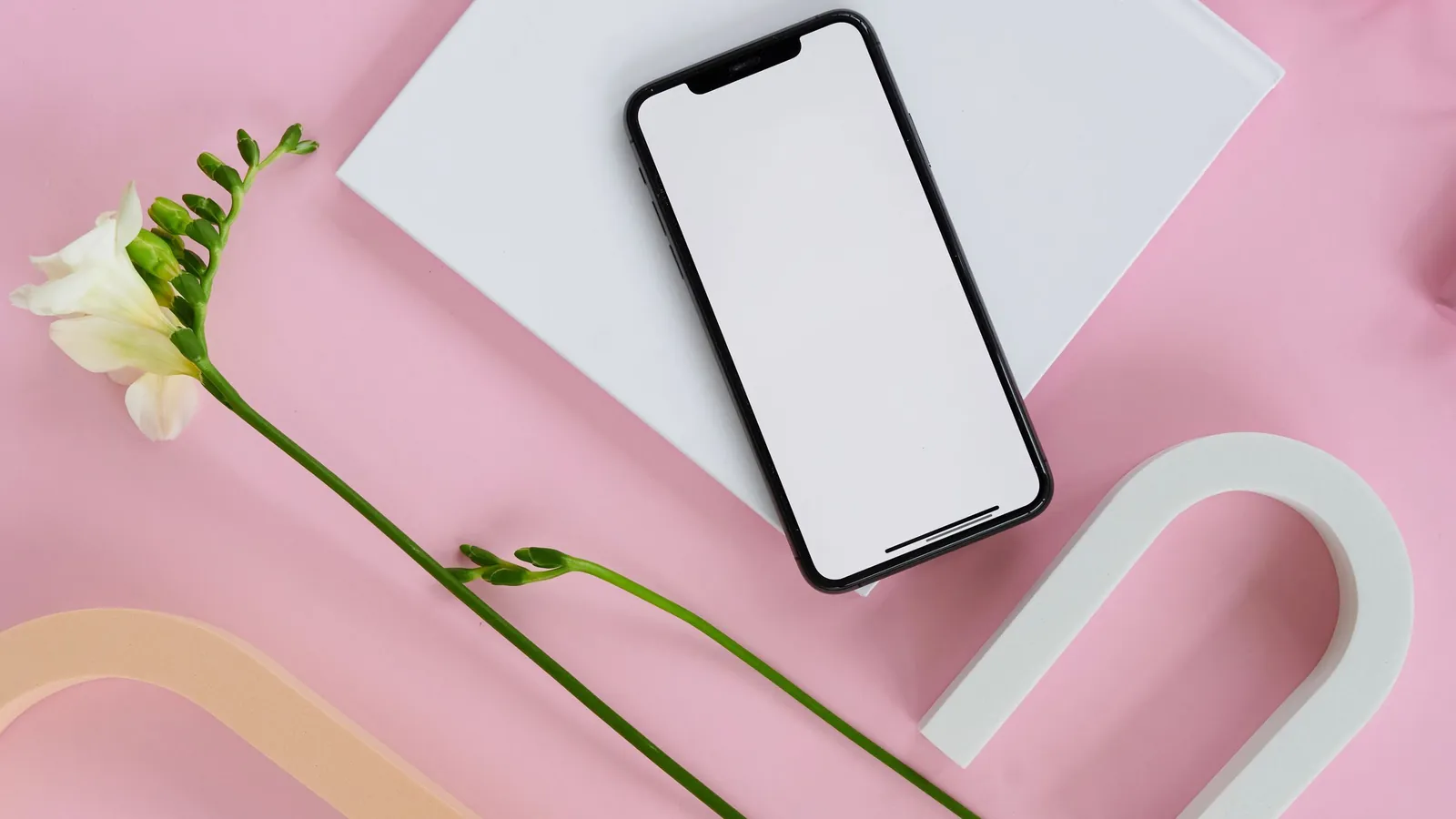 Smartphone with a blank screen on a pink flat-lay backdrop with a white freesia and pastel shapes