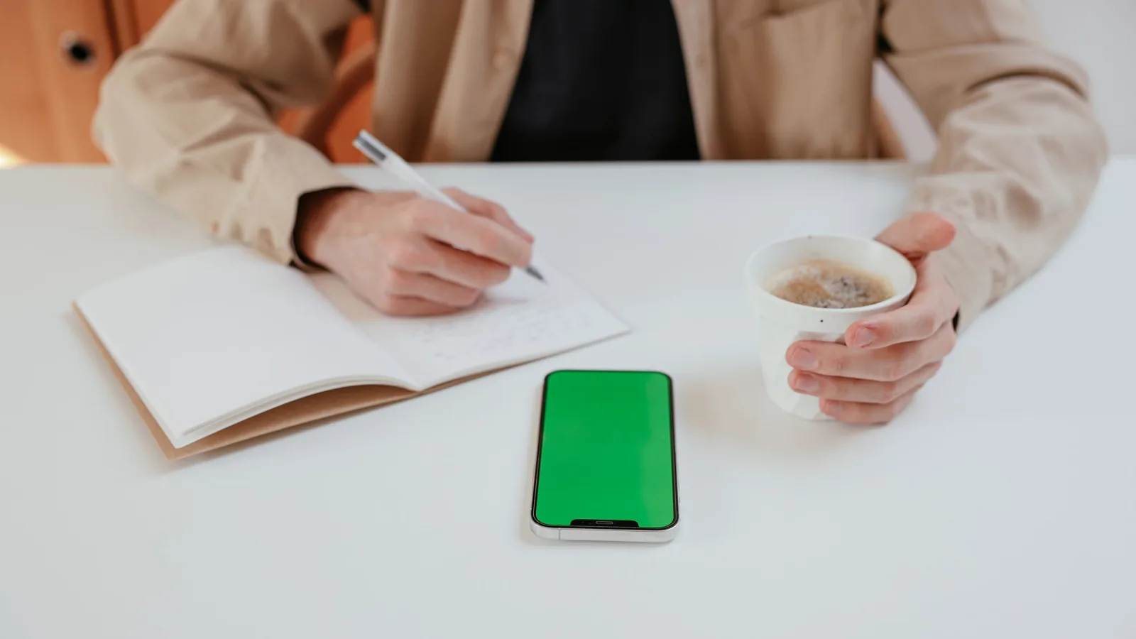 Person writing in a notebook at a white desk, holding a cup of coffee